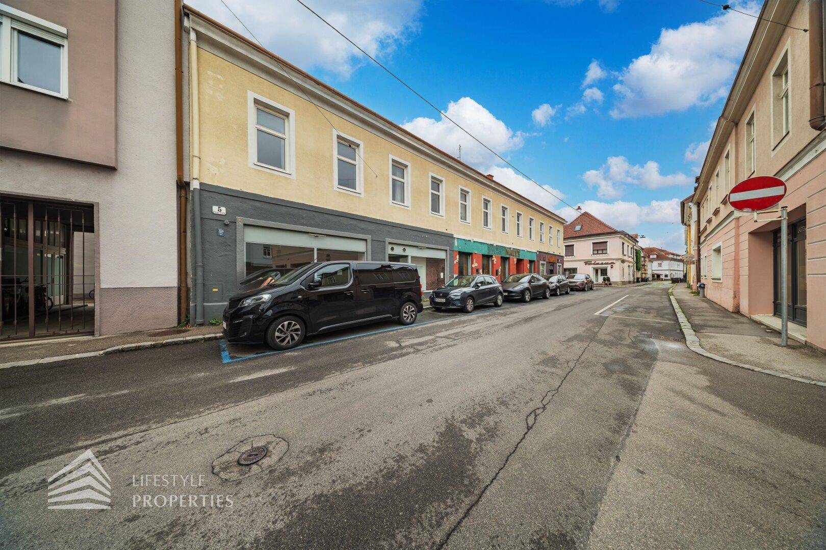 Lange Reihe von Gebäuden entlang einer Straße mit geparkten Autos und einem blauen Himmel.
