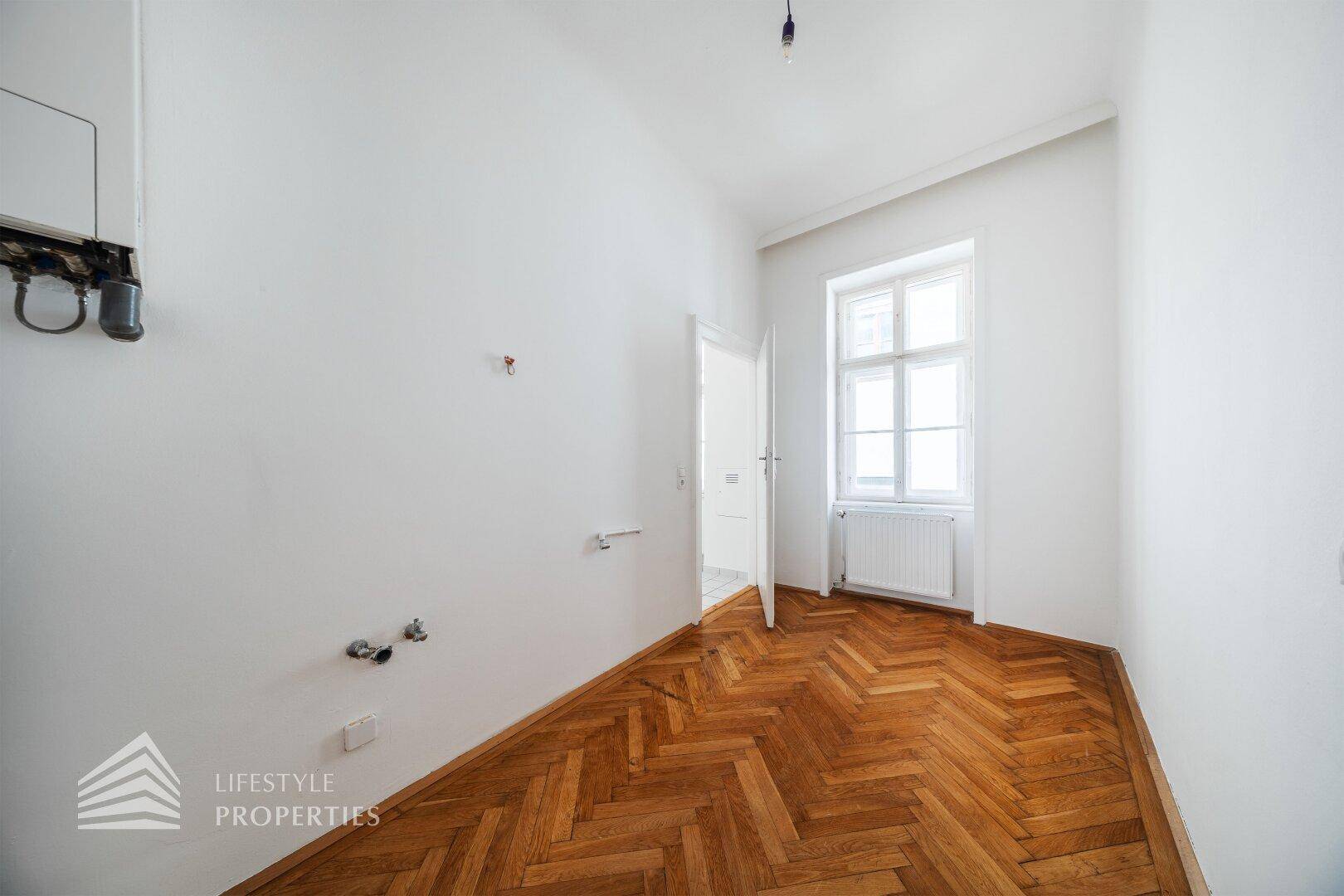Raum mit Fischgrätparkett, Fenster und Anschlüssen für Installationen, möglicherweise eine Küche.