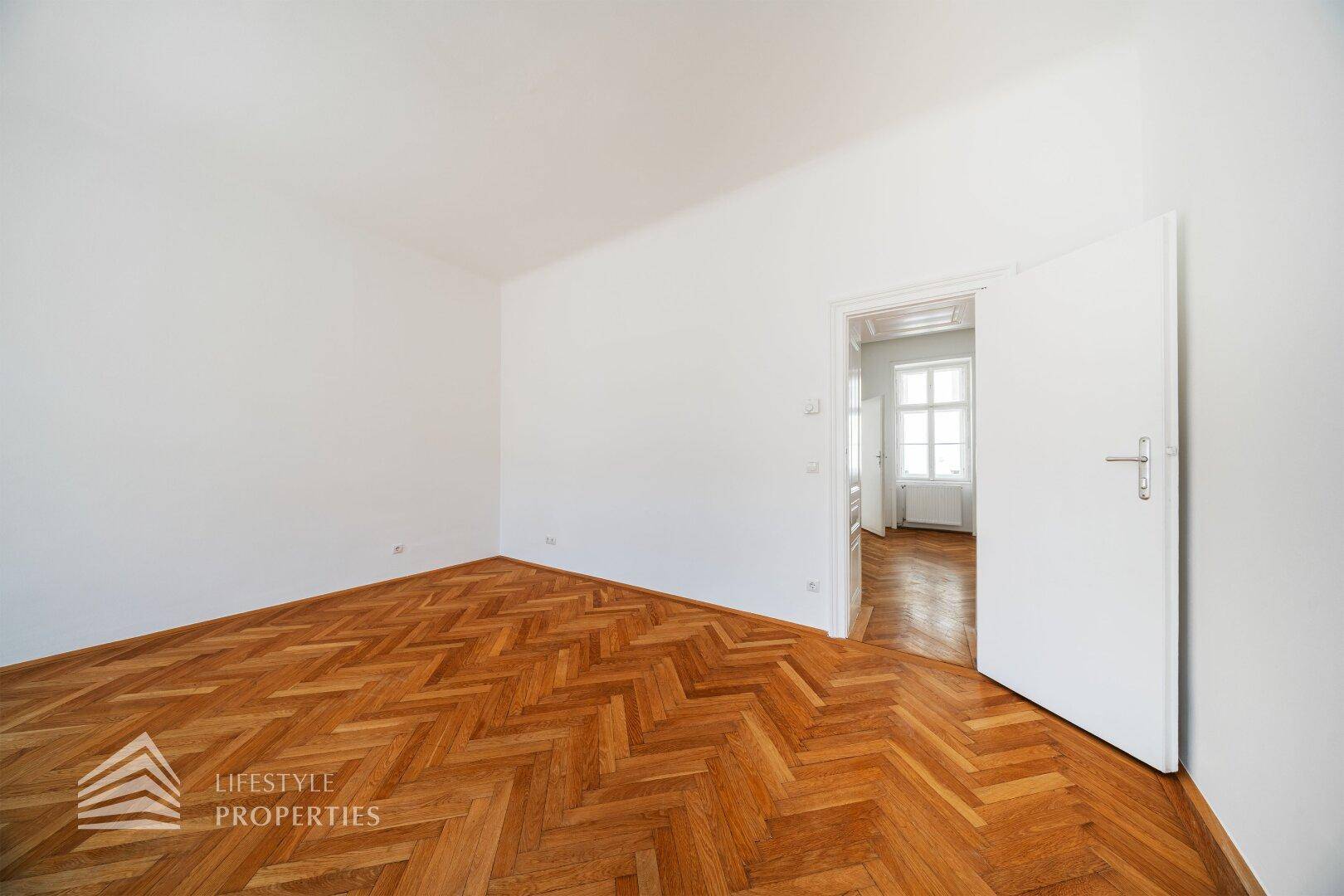 Leeres Zimmer mit Fischgrätparkett und offener Tür zu einem angrenzenden Bereich.