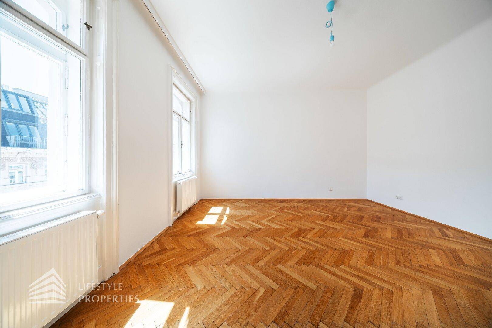 Geräumiges Zimmer mit klassischem Fischgrätparkett und viel Tageslicht durch die Fenster.