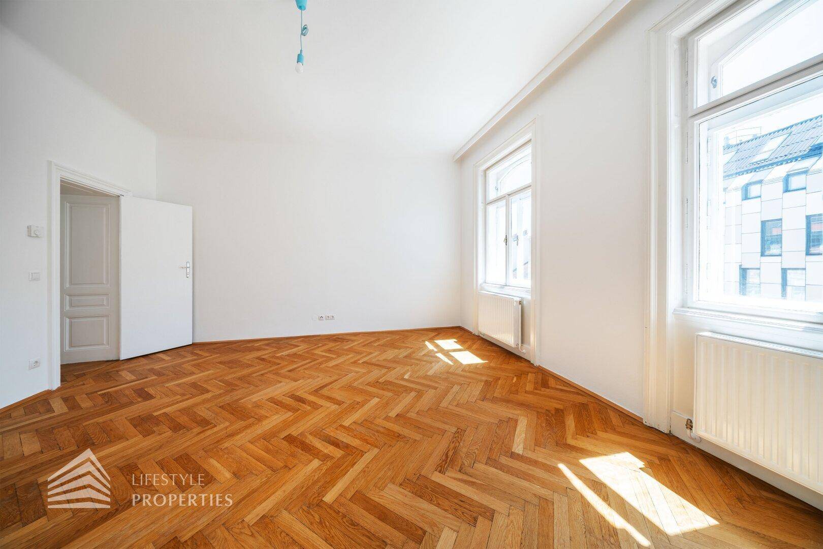 Heller Raum mit Fischgrätparkett und einer Tür, die zu einem weiteren Zimmer führt.