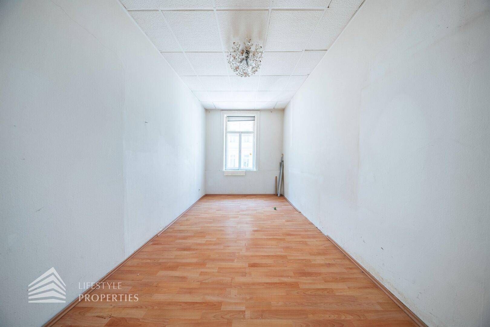 Langer, schmaler Raum mit Laminatboden, einem Fenster und Deckenleuchte.
