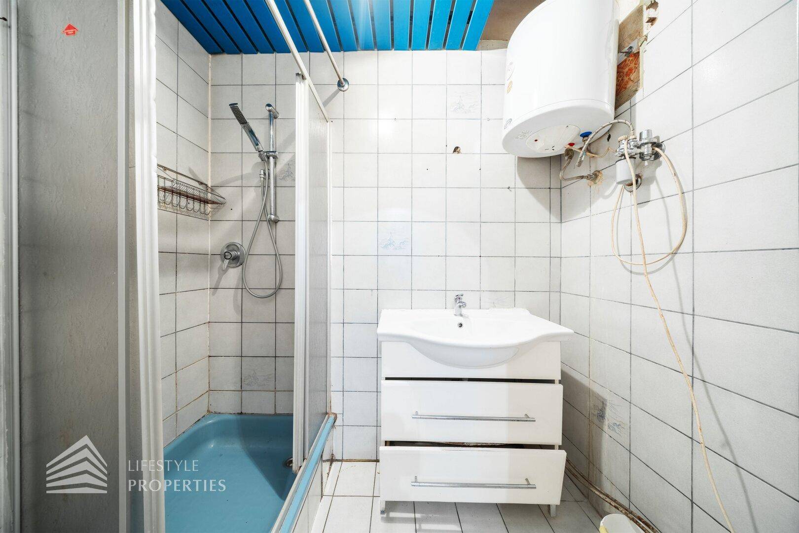 Kleines Badezimmer mit Dusche, Waschbecken und Boiler, gefliest in Weiß und Blau.