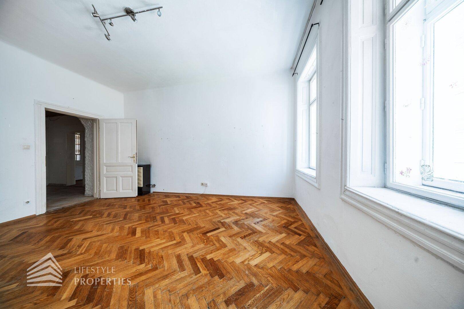 Geräumiges Zimmer mit Fischgrätparkett, hohen Decken und einem Ofen, bereit zur Renovierung.