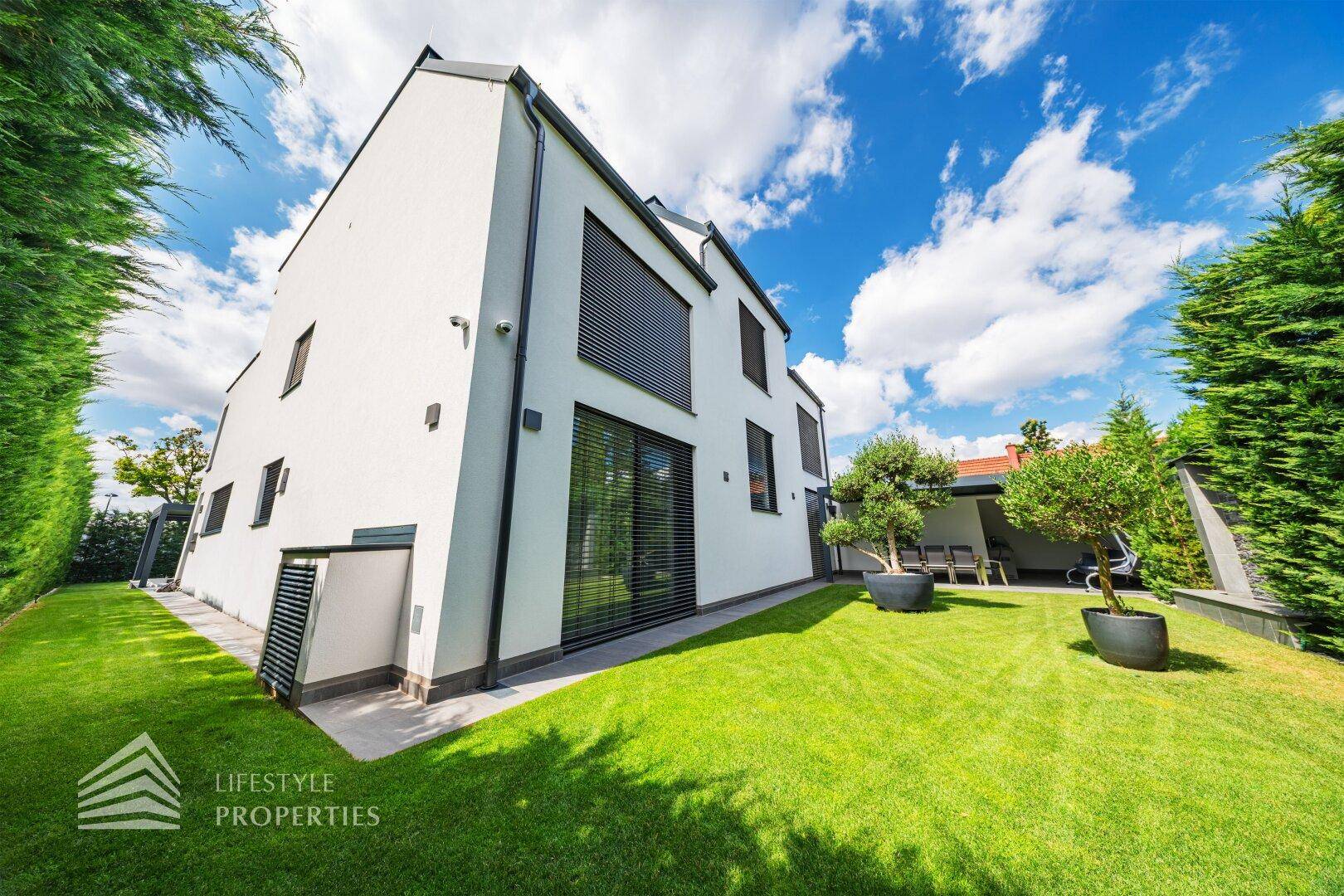 Moderne Villa mit großem, gepflegtem Garten und dekorativen Olivenbäumen in Töpfen.