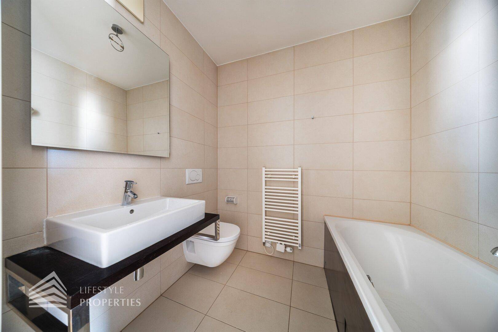 Elegantes Badezimmer mit beigen Fliesen, Waschtisch, wandhängendem WC und einer Badewanne.