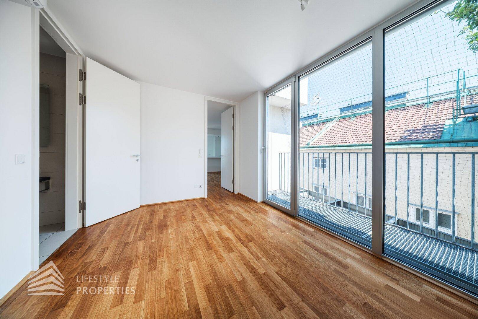 Schlafzimmer mit Holzboden und großen Fenstern, die auf einen kleinen Balkon blicken.
