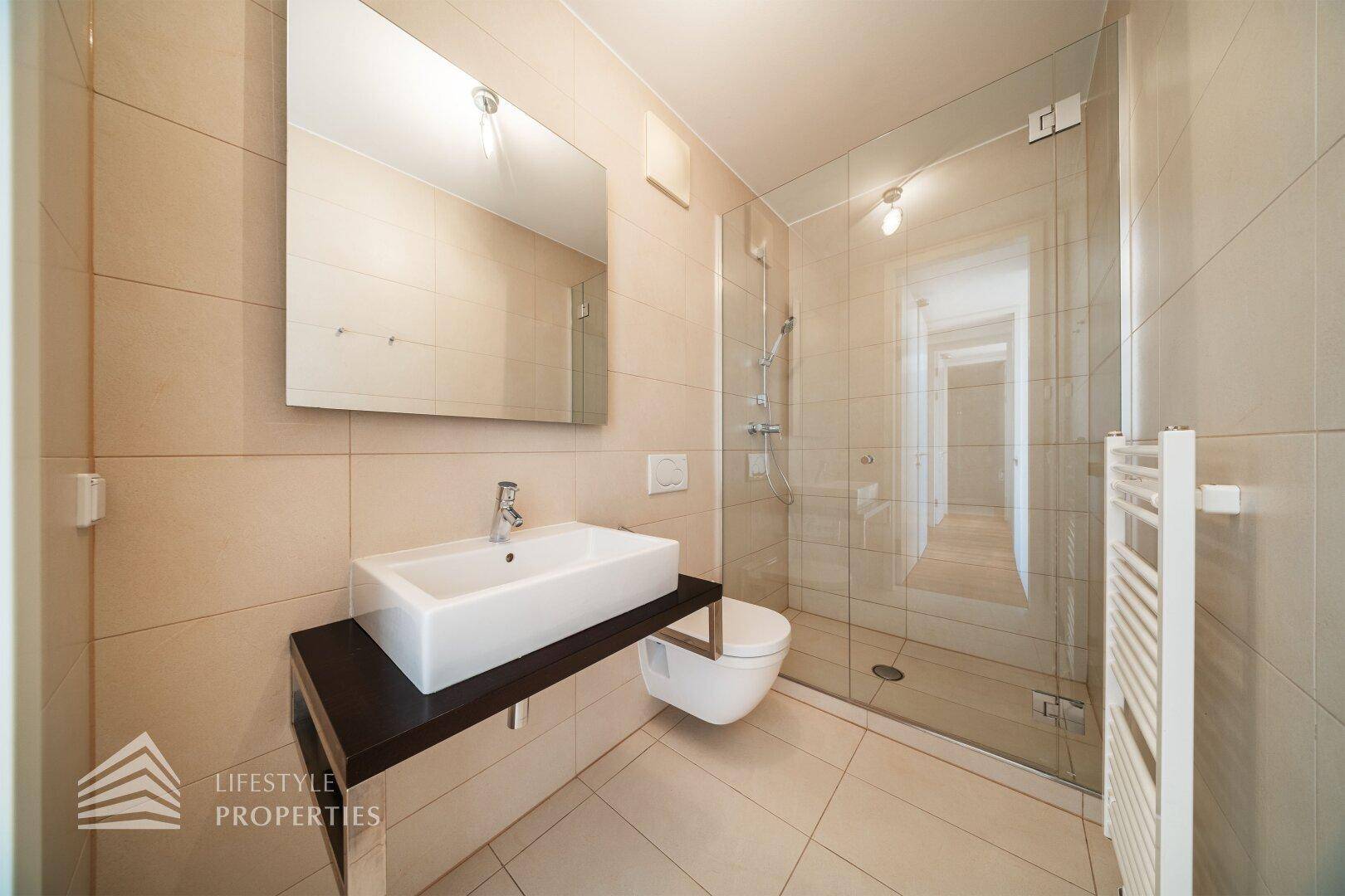 Modernes Badezimmer mit beigen Fliesen, Waschtisch, wandhängendem WC und begehbarer Dusche.