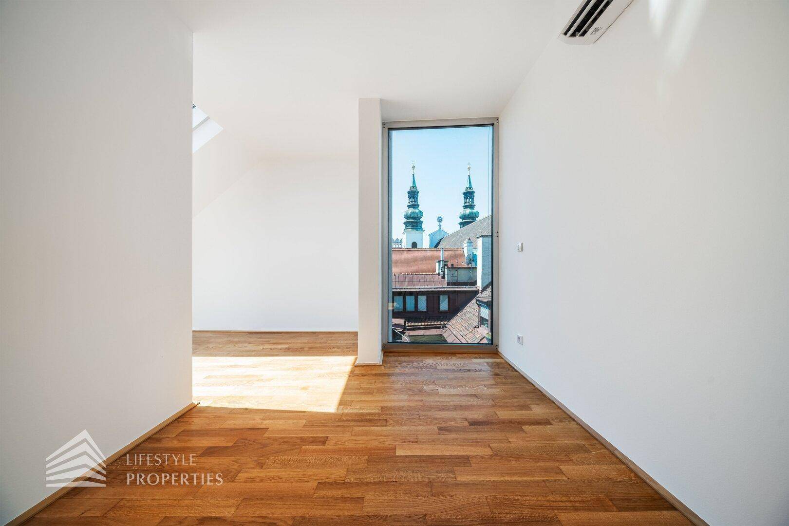Heller Raum mit Holzboden, weißen Wänden und Blick auf Kirchtürme. Eine Klimaanlage ist installiert.