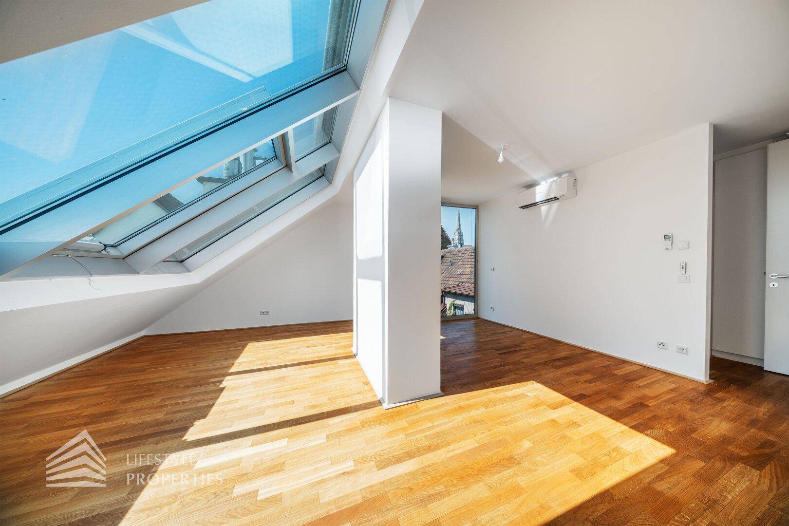 Helles Dachgeschosszimmer mit Holzboden, Dachfenstern und einem vertikalen Fenster mit Ausblick.