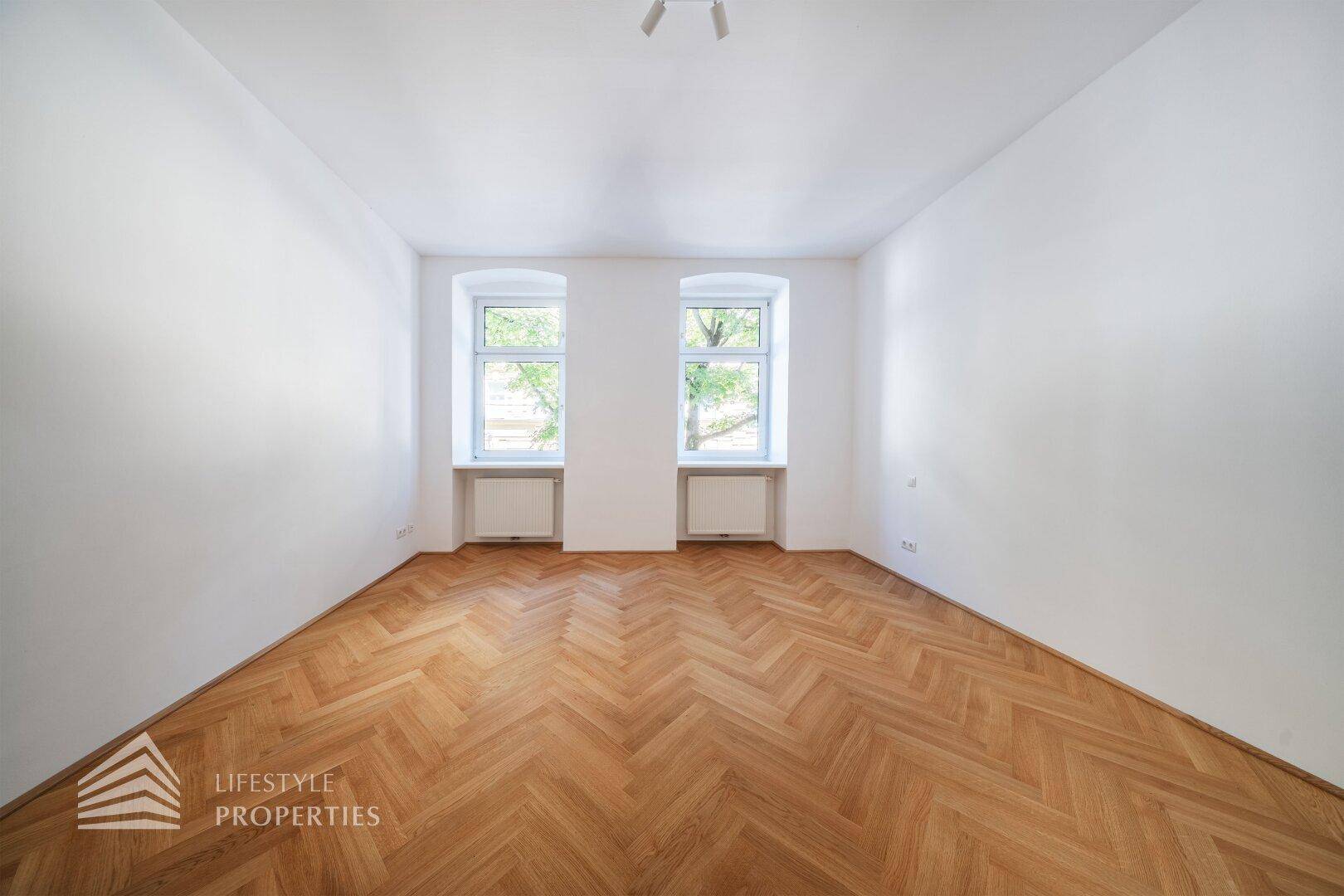 Leeres Zimmer mit hellem Fischgrätparkett und zwei Fenstern, bereit zur individuellen Gestaltung.