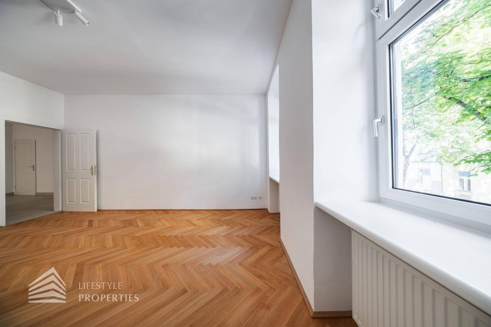 Heller Raum mit Fischgrätparkett und einer Tür, die zu einem weiteren Zimmer führt.