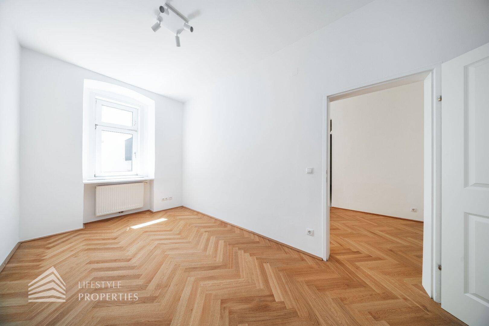 Geräumiges Zimmer mit Fischgrätparkett, einem Fenster und einer Tür zu einem angrenzenden Raum.