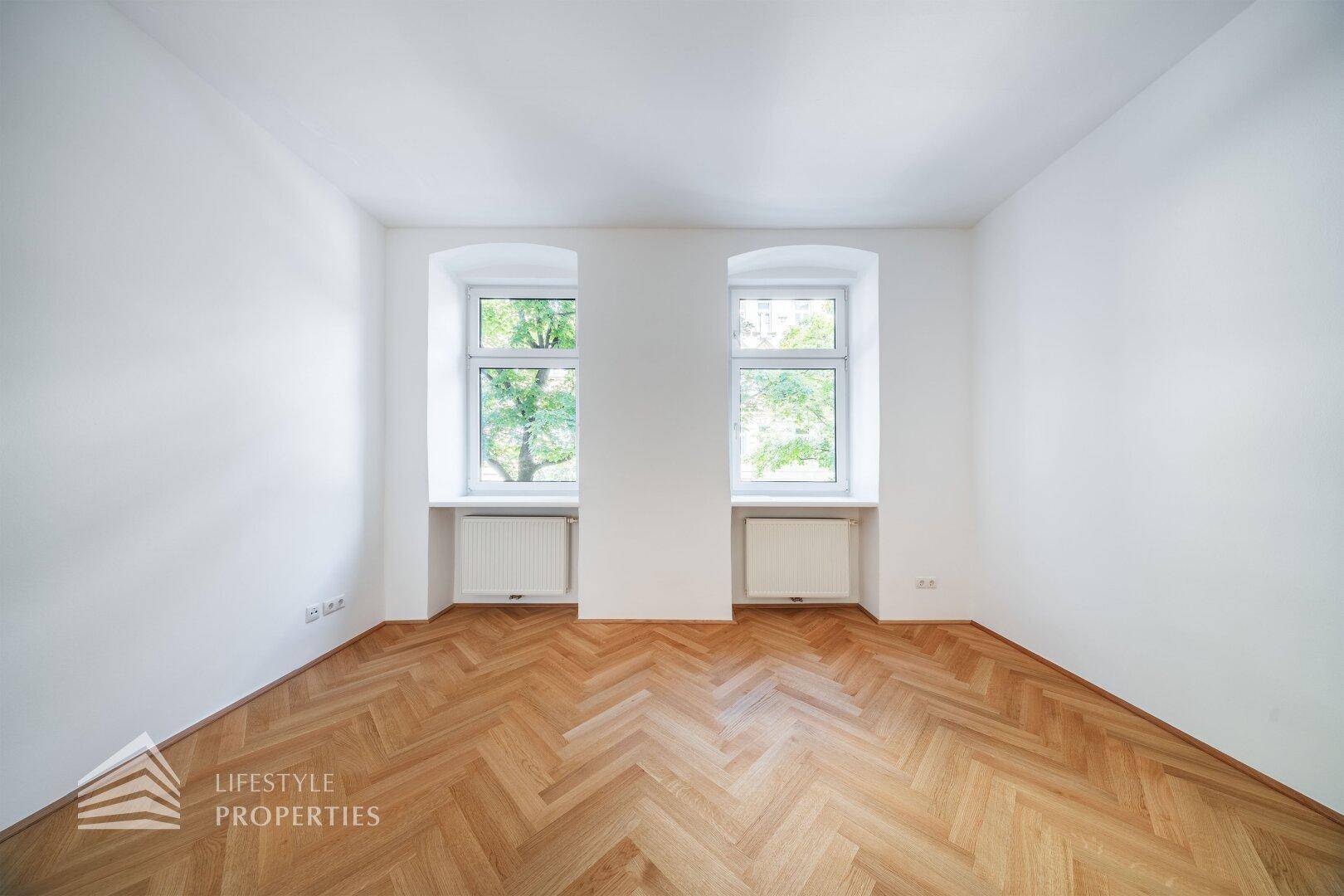 Geräumiges Zimmer mit klassischem Fischgrätparkett und zwei großen Fenstern mit Blick ins Grüne.