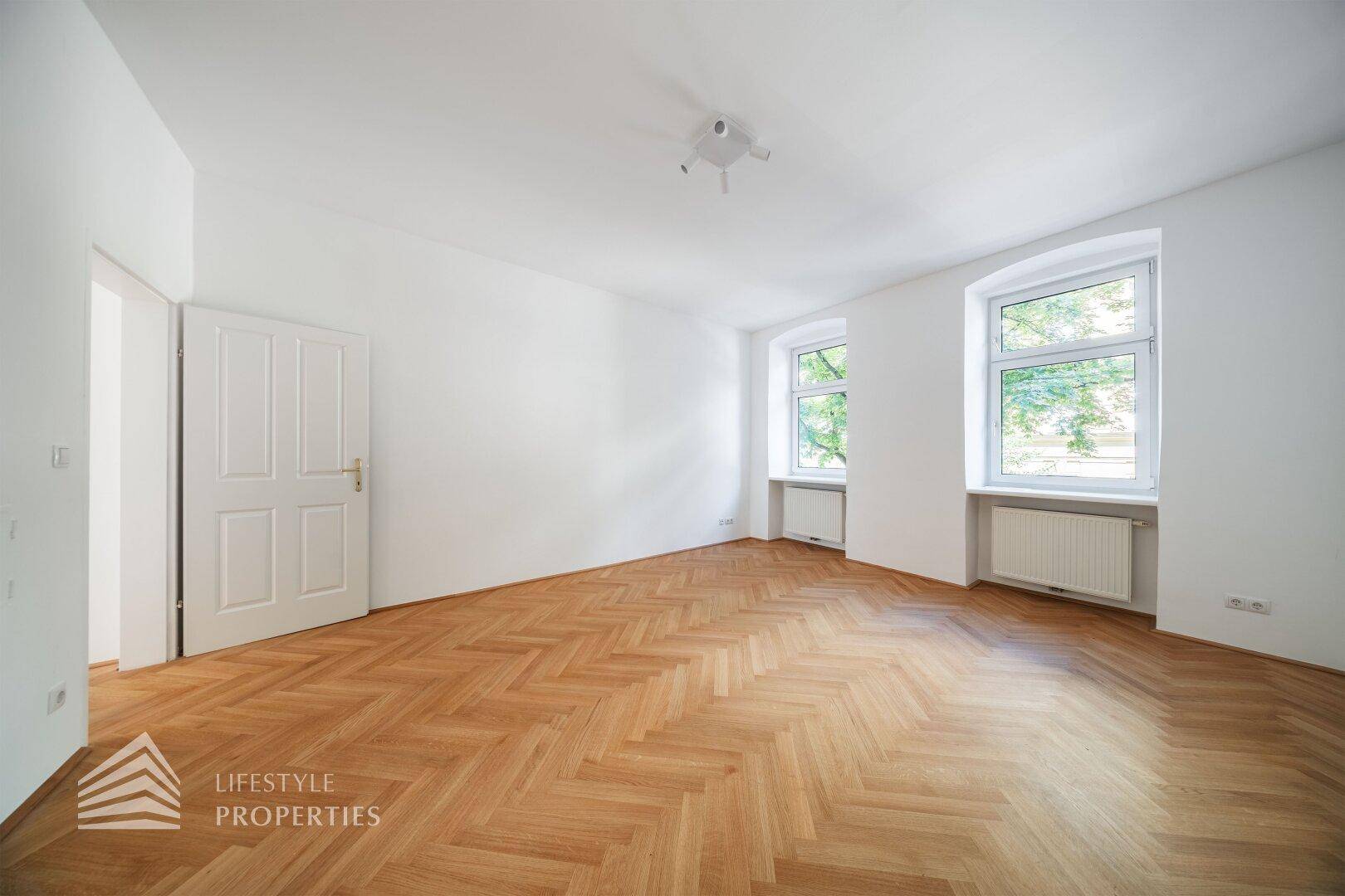 Großes, helles Zimmer mit Fischgrätparkett, zwei Fenstern und einer weißen Holztür.