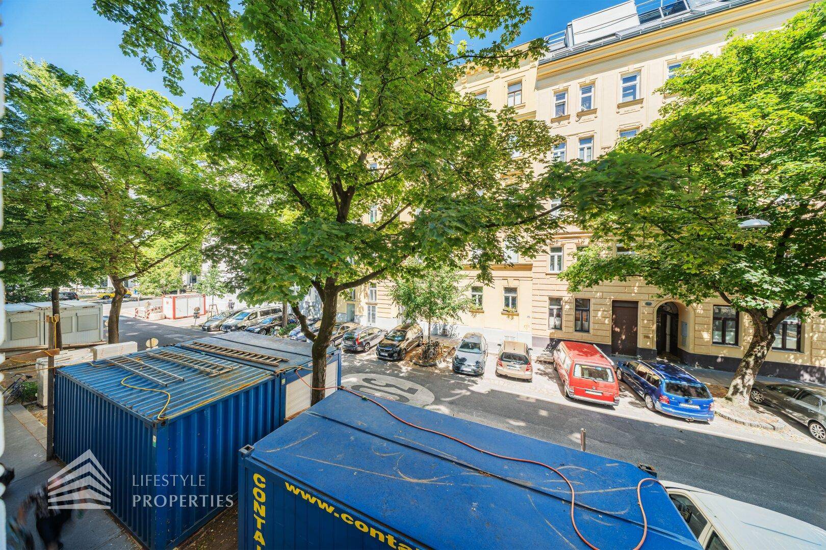 Blick von oben auf eine belebte Straße mit parkenden Autos und grünen Bäumen.