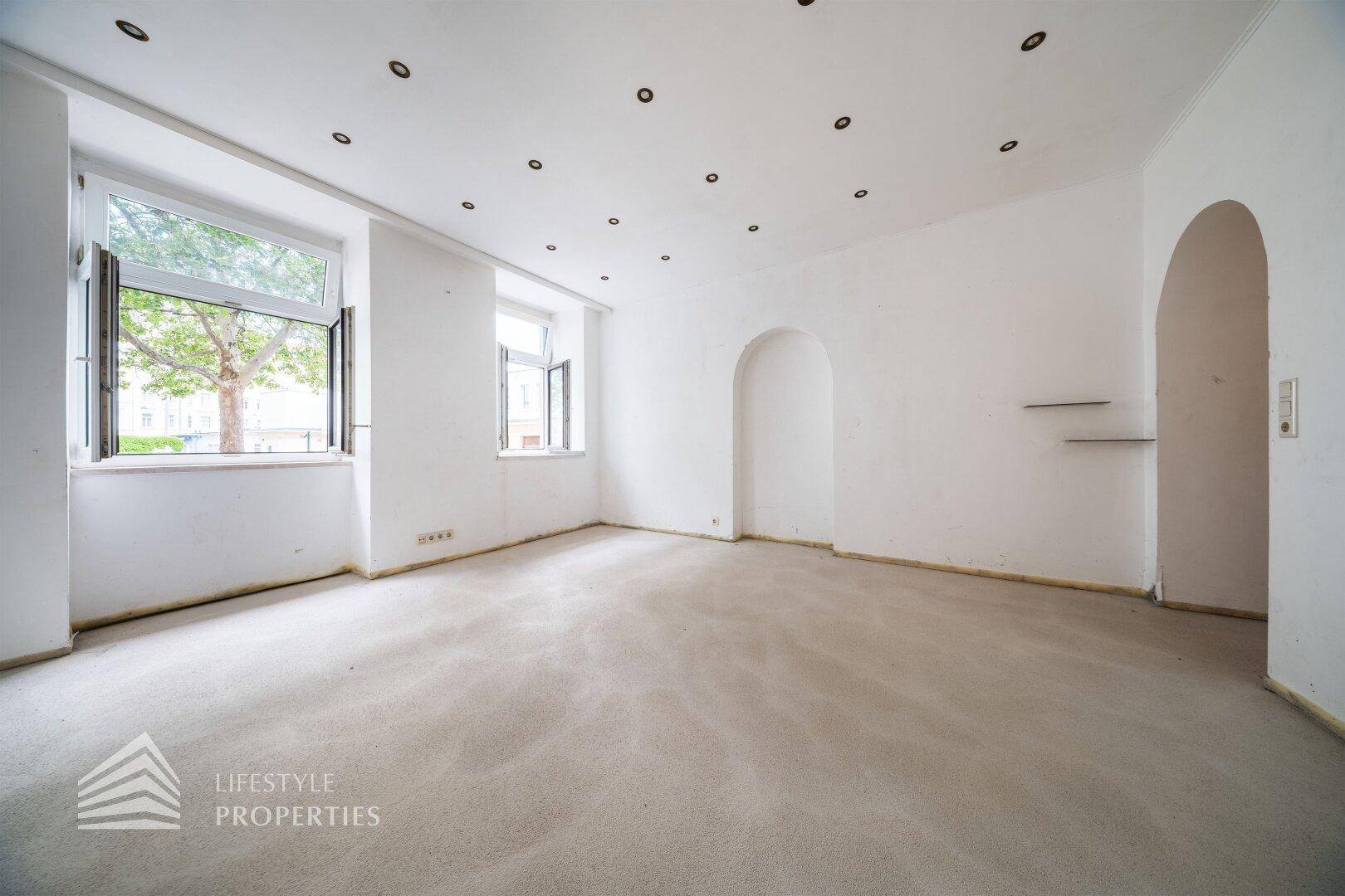 Geräumiger, leerer Wohnraum mit weißen Wänden, Teppichboden, Einbaustrahlern und zwei Fenstern mit Blick ins Grüne.