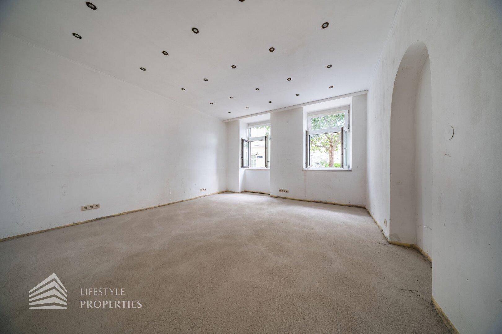 Heller, unmöblierter Wohnraum mit Teppichboden, weißen Wänden, Einbaustrahlern und zwei Fenstern mit Tageslicht.