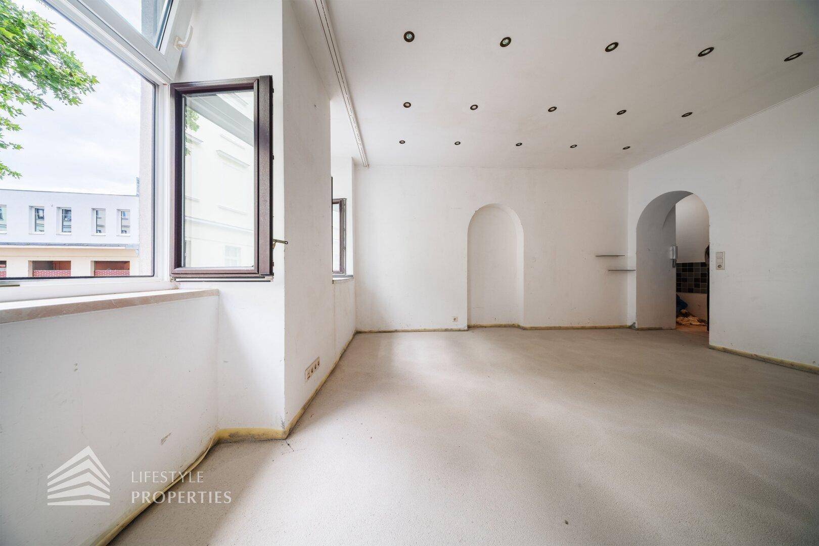 Geräumiger, heller Wohnraum mit Teppichboden, weißen Wänden, Einbaustrahlern und großen Fenstern mit Tageslicht.