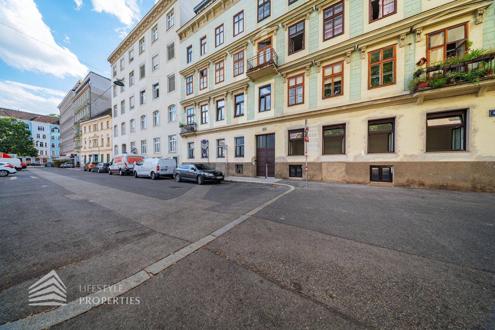 Ansicht einer mehrstöckigen Wohnhausfassade an einer Straße mit parkenden Fahrzeugen und Balkonen.