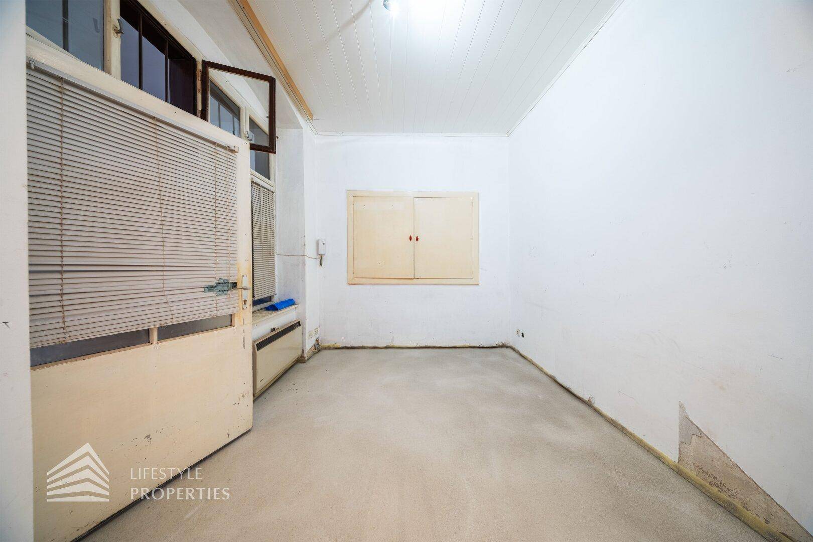 Heller, unmöblierter Raum mit großen Fenstern, Jalousien und einer schlichten Wandgestaltung, bereit zur Renovierung.