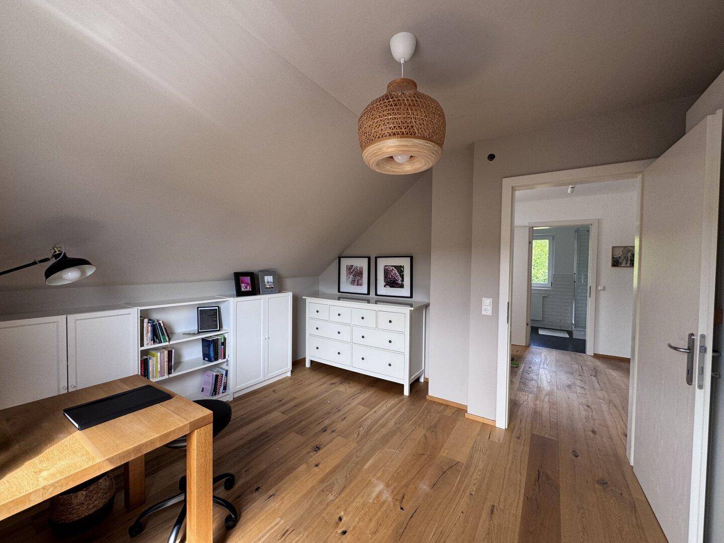 Helles Arbeitszimmer mit Holzboden, Schreibtisch, Regalen und Dachschräge.