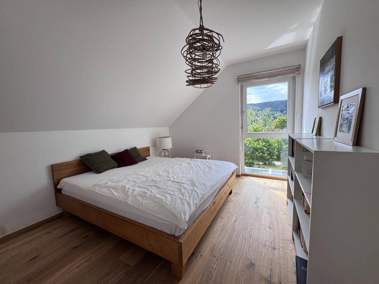 Helles Schlafzimmer mit Holzbett, Holzboden, Bücherregal und weitem Ausblick.