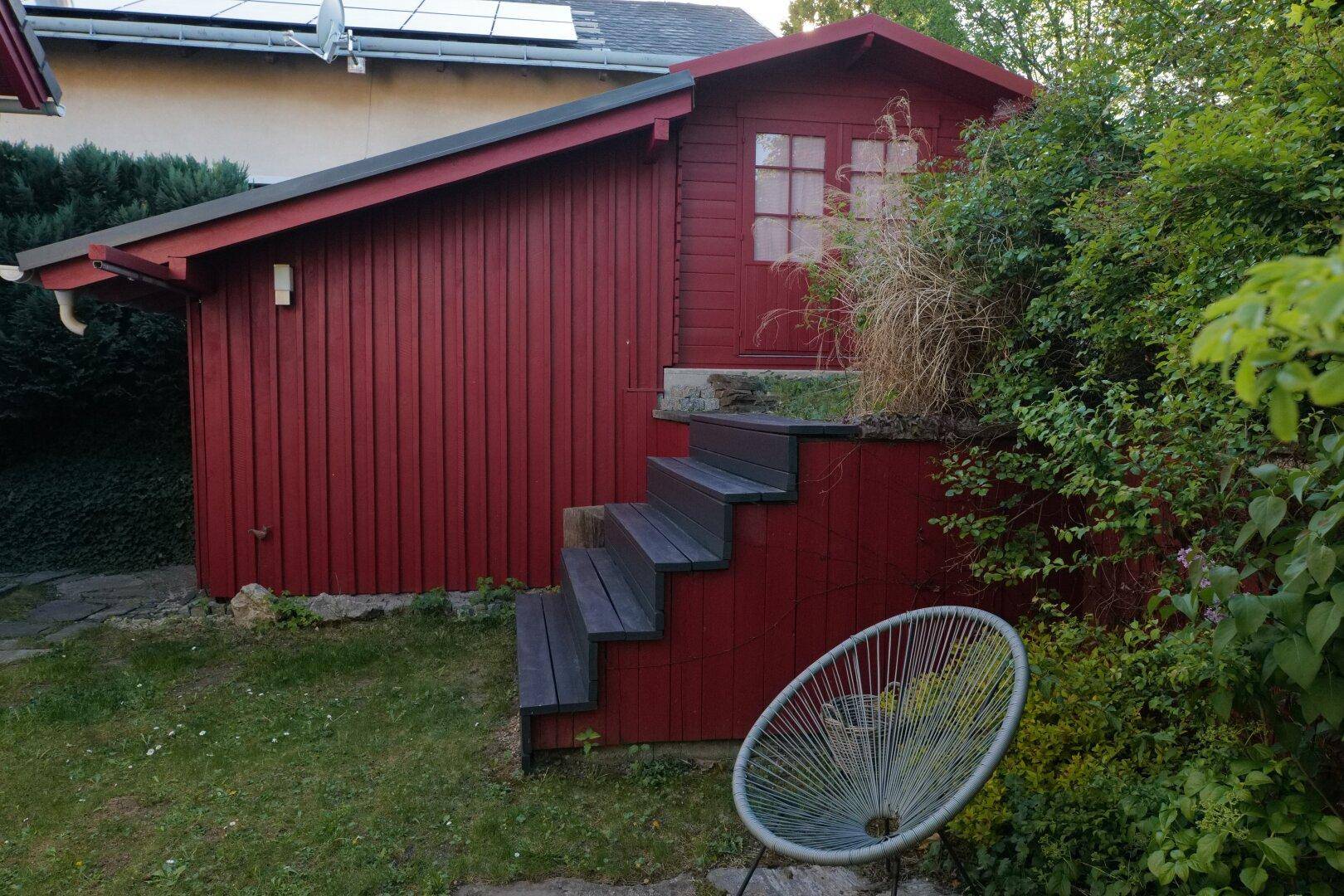 Rotes Gartenhaus mit Holzstufen und umgebender Bepflanzung im Garten.
