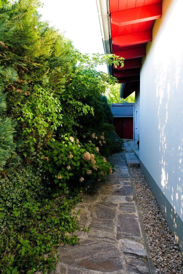 Steinweg führt entlang der Hauswand, gesäumt von dichtem Grün und blühenden Sträuchern.