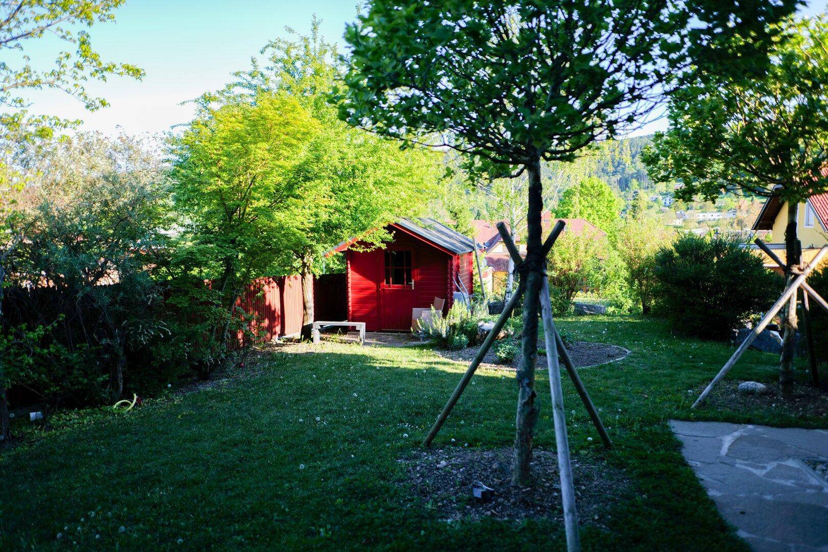Gepflegter Garten mit Rasenfläche, jungen Bäumen und einem charmanten roten Gartenhaus.