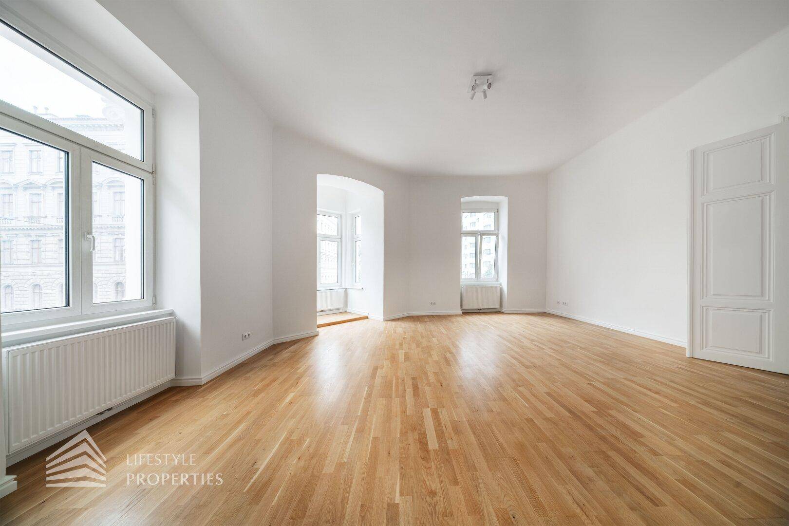 Lichtdurchflutetes Wohnzimmer mit klassischem Parkettboden und mehreren Fenstern, ideal für helle Einrichtung.