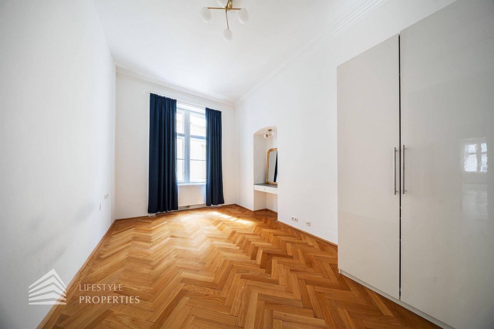 Schlafzimmer mit Fischgrätparkett, einem großen Fenster und einem modernen weißen Kleiderschrank.