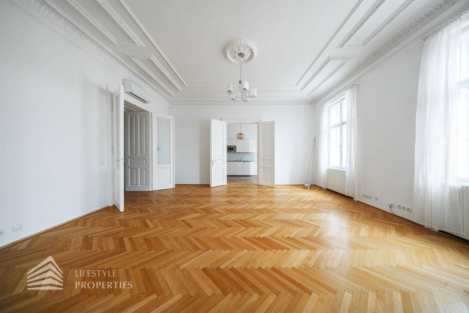 Helles Wohnzimmer mit klassischem Fischgrätparkett und hohen Decken, mit Blick auf die angrenzende Küche.