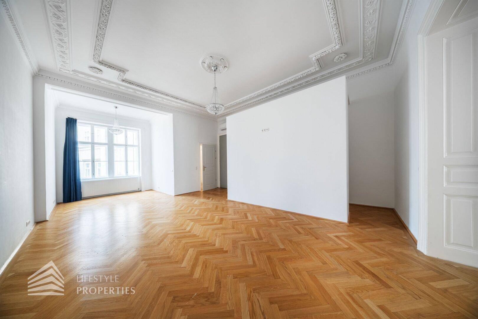 Geräumiges Wohnzimmer mit Fischgrätparkett, hohen Decken und einem großen Fenster mit blauen Vorhängen.