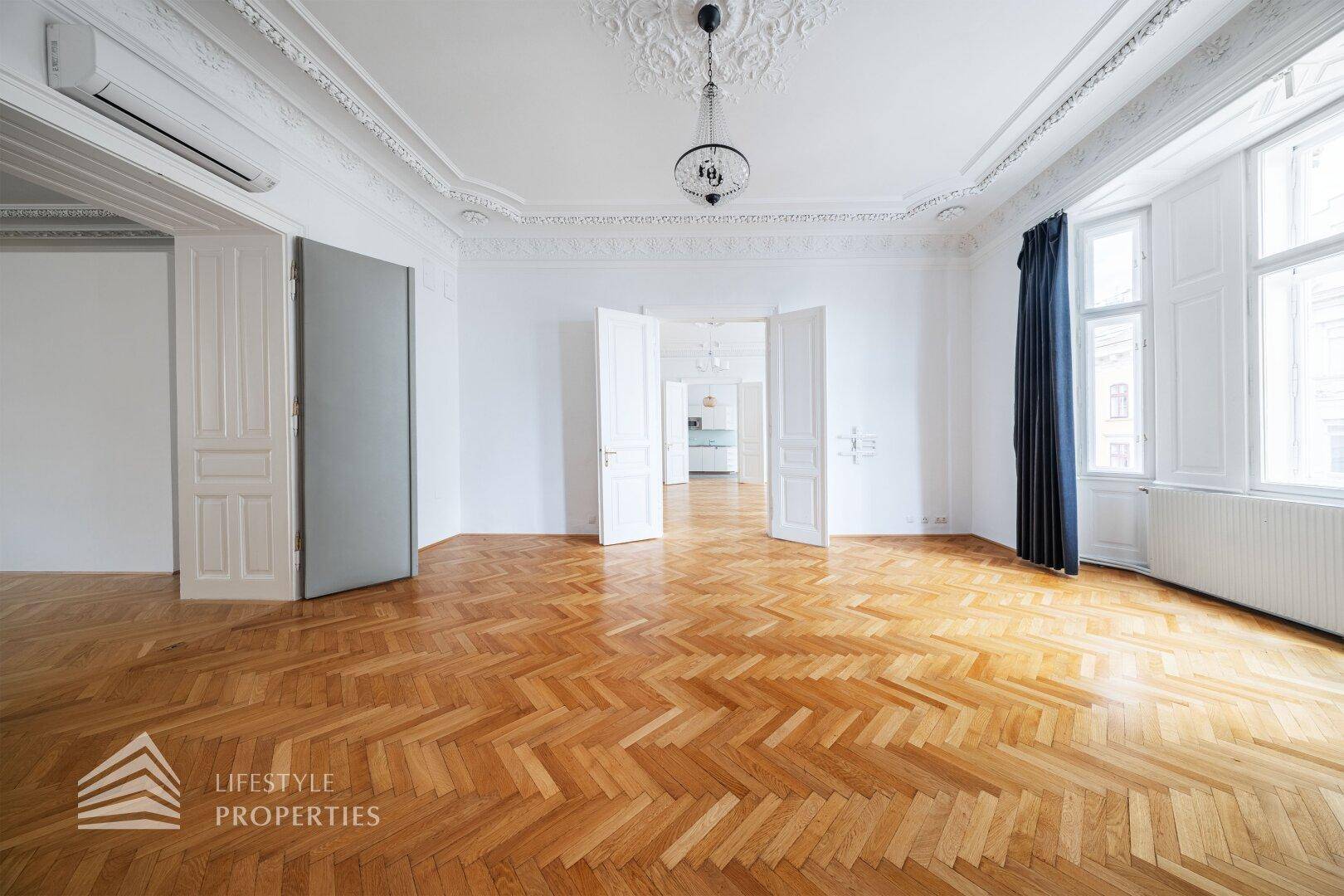 Elegantes Wohnzimmer mit Fischgrätparkett, Stuckdecken und Klimaanlage, mit Blick in weitere Räume.