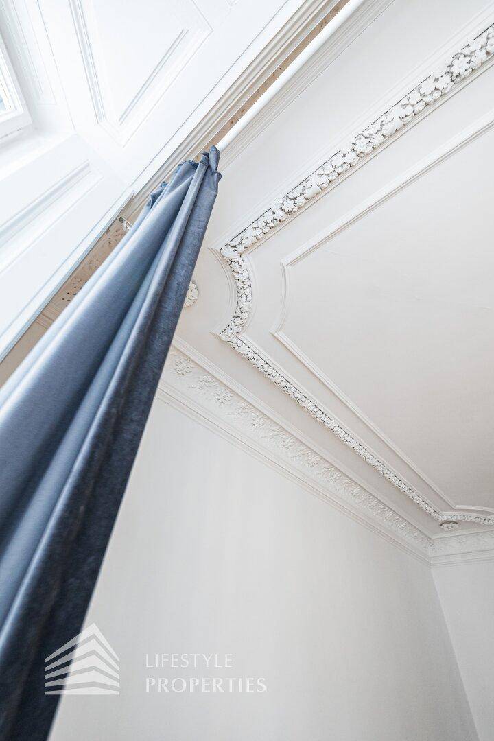 Detailaufnahme einer hohen Decke mit Stuckverzierungen und einem blauen Vorhang am Fenster.