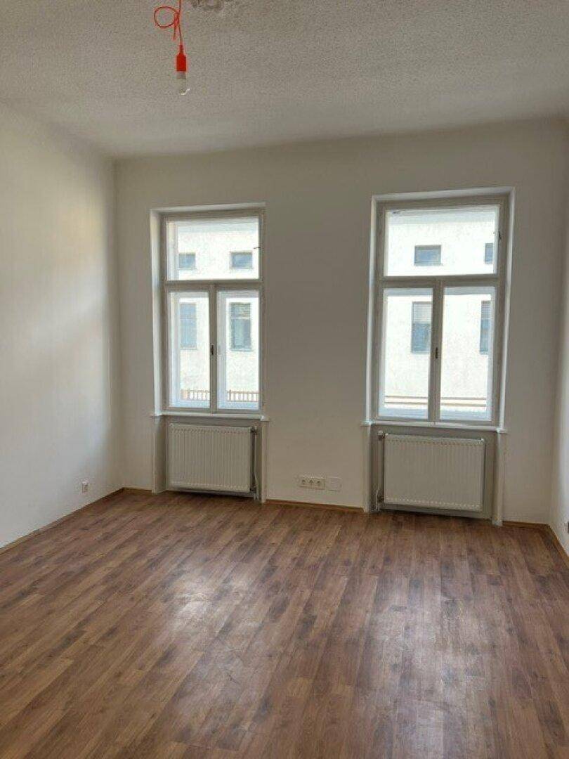 Heller, leerer Raum mit zwei Fenstern, Laminatboden und weiß gestrichenen Wänden.