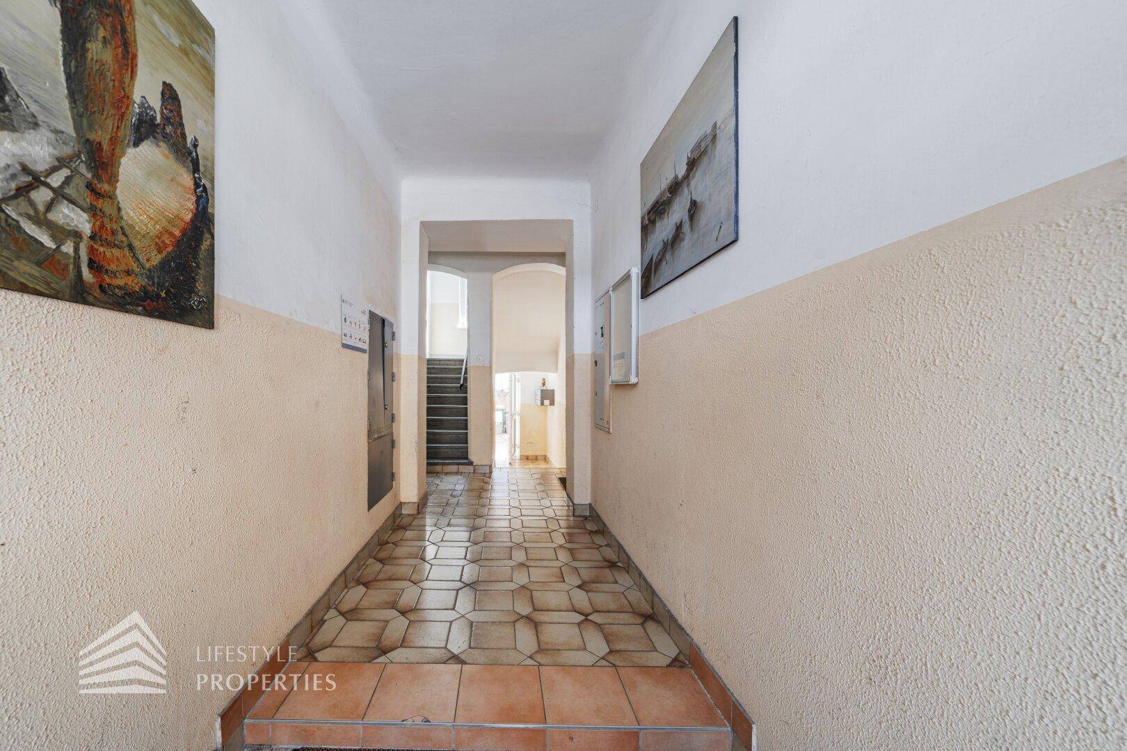Langer Flur mit zweifarbigen Wänden und gemusterten Fliesenboden, der zu einer Treppe führt.