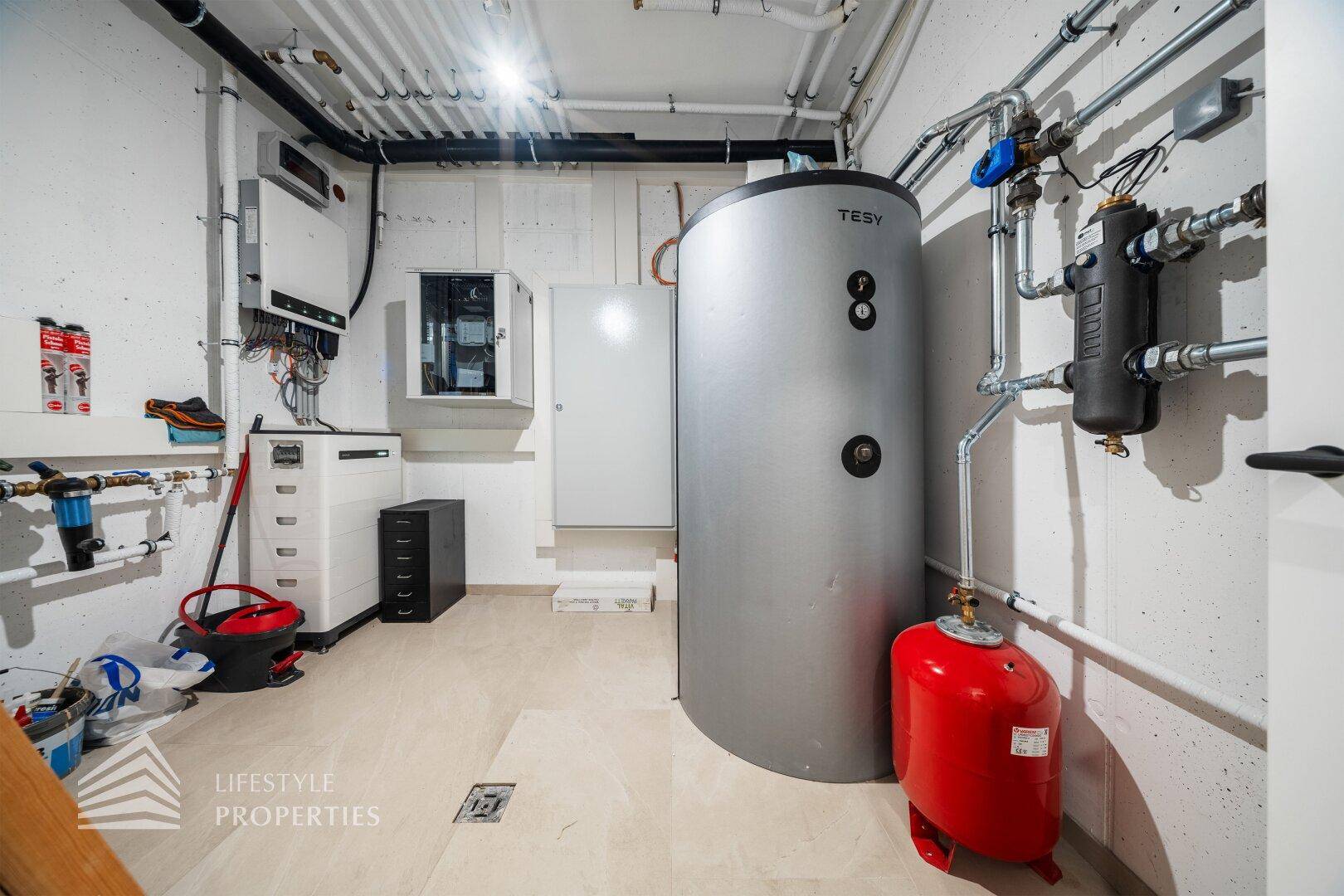Technikraum mit Warmwasserspeicher, Heizungsanlage und zahlreichen Rohrleitungen.