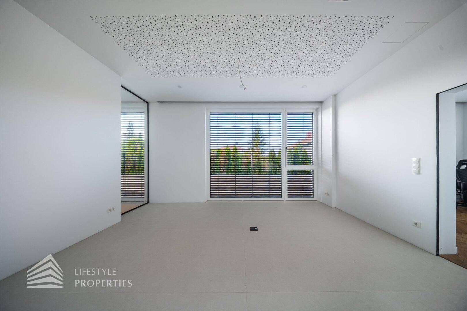 Heller, leerer Raum mit großen Fenstern, Jalousien und modernem, minimalistischem Design.