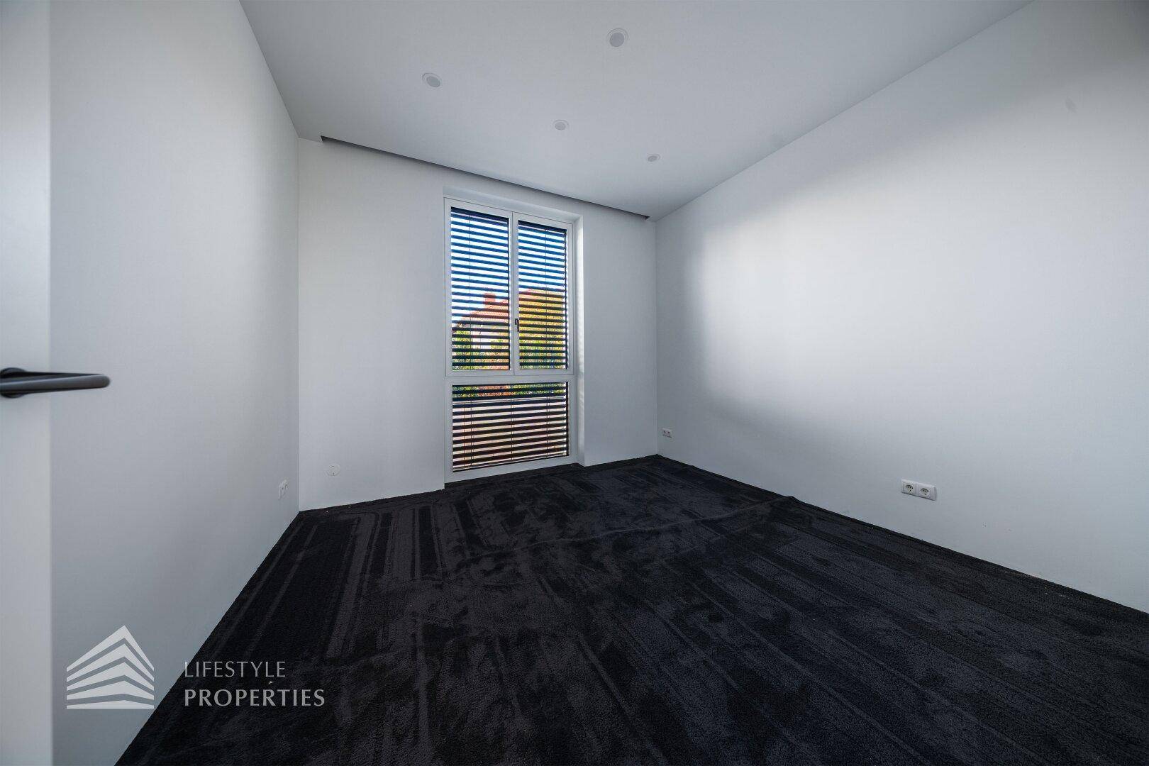 Leeres Zimmer mit dunklem Teppichboden, weißen Wänden und einem Fenster mit Jalousien.