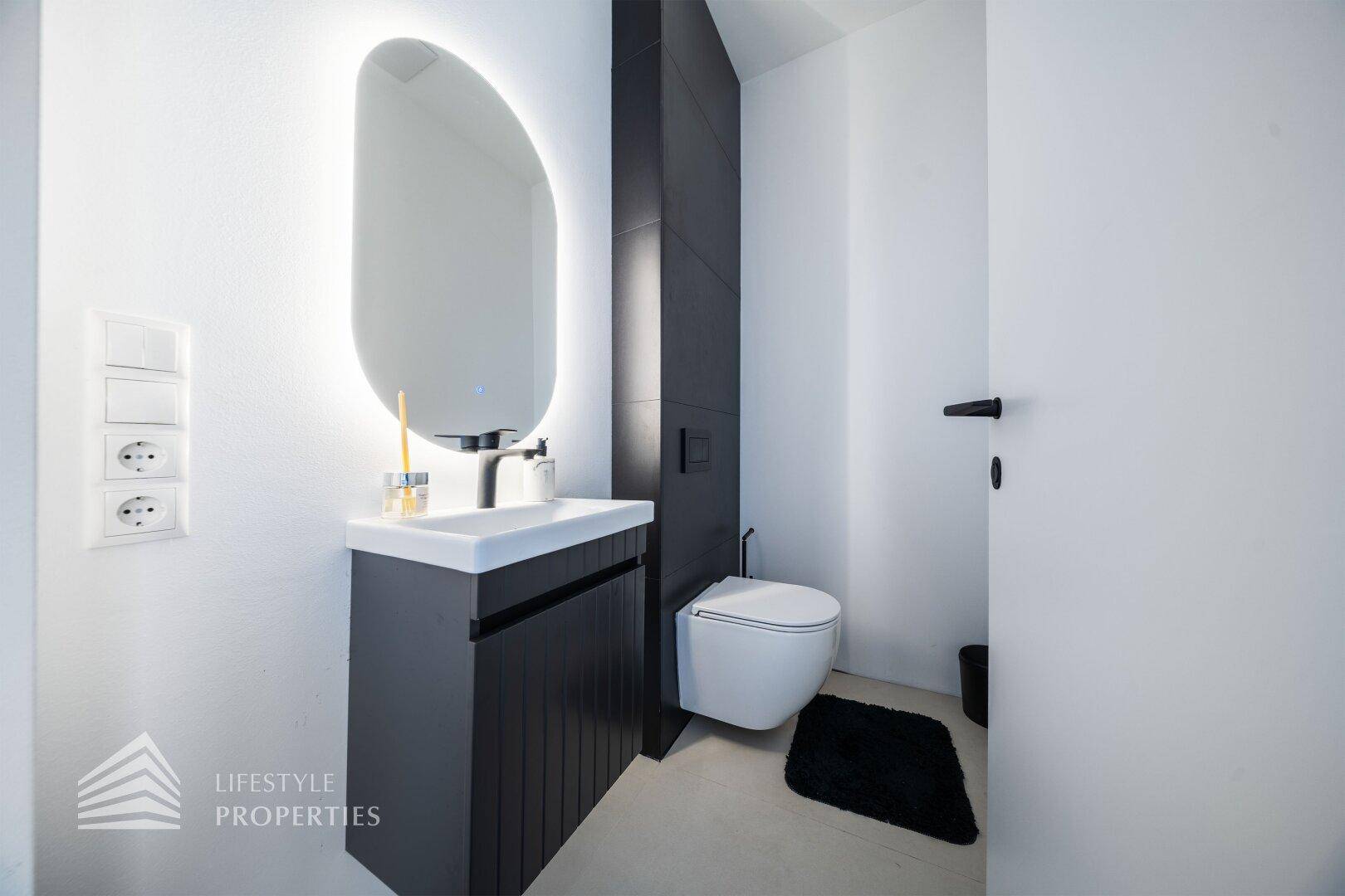 Modernes Gäste-WC mit wandhängender Toilette, beleuchtetem Spiegel und dunkler Akzentwand.