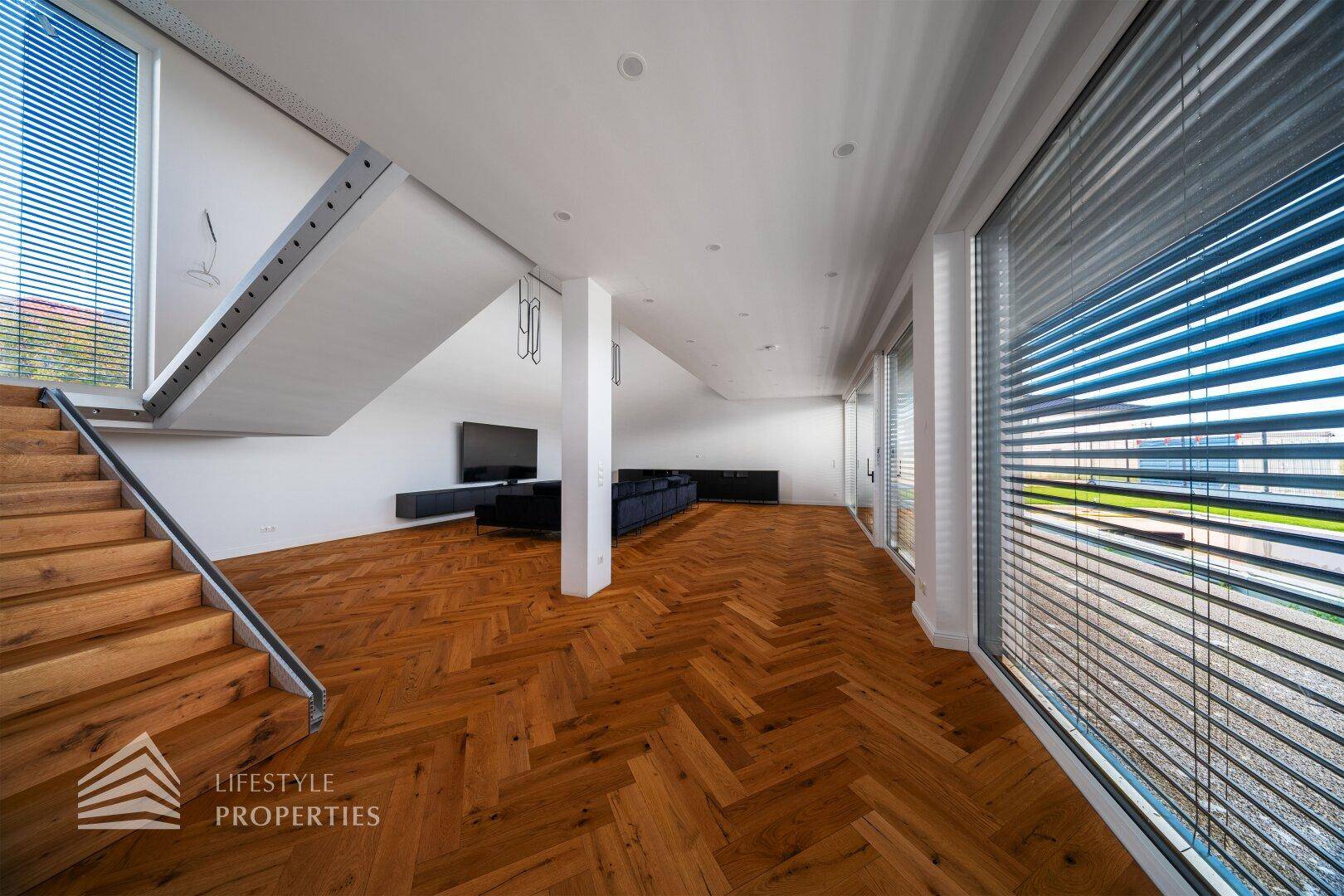 Geräumiges Wohnzimmer mit Fischgrätparkett, Holztreppe und großen Fenstern mit Jalousien.