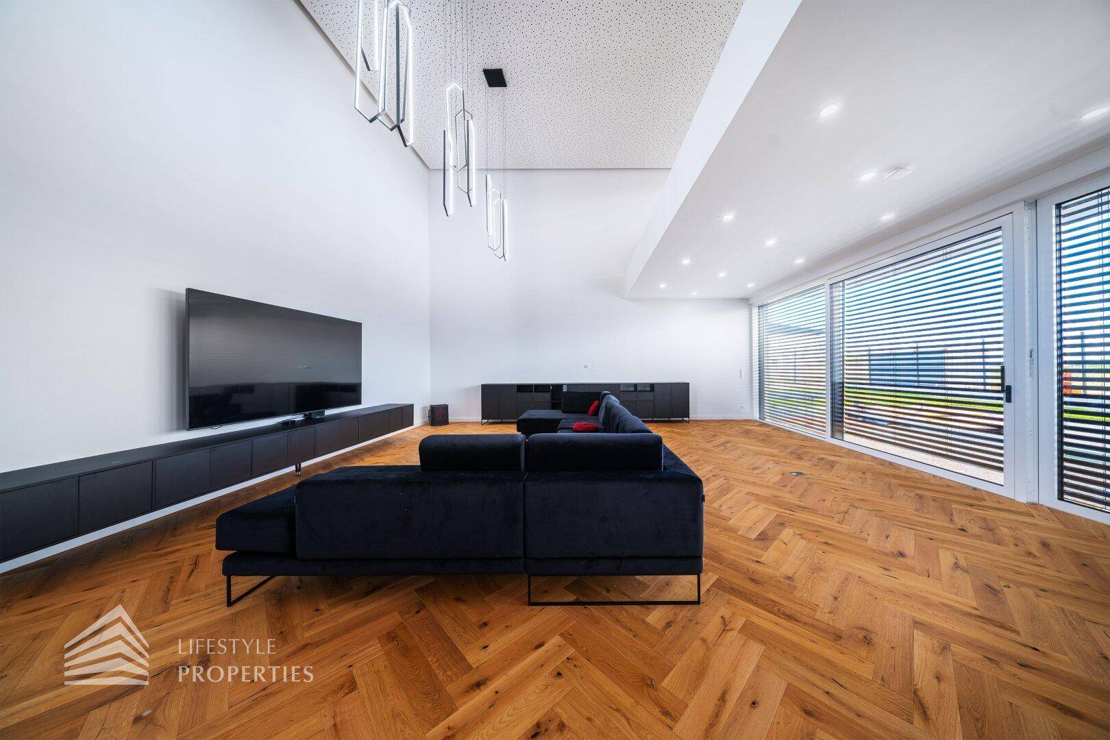 Modernes Wohnzimmer mit dunklem Sofa, großem Fernseher und Fischgrätparkettboden.
