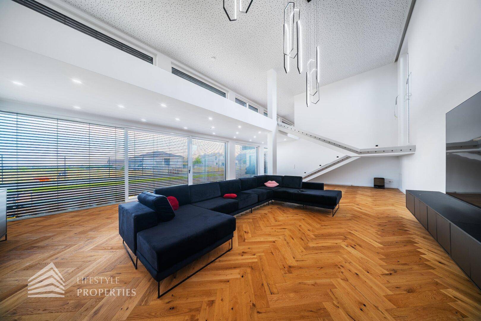 Großzügiges Wohnzimmer mit hohen Decken, dunklem Sofa und Fischgrätparkettboden.