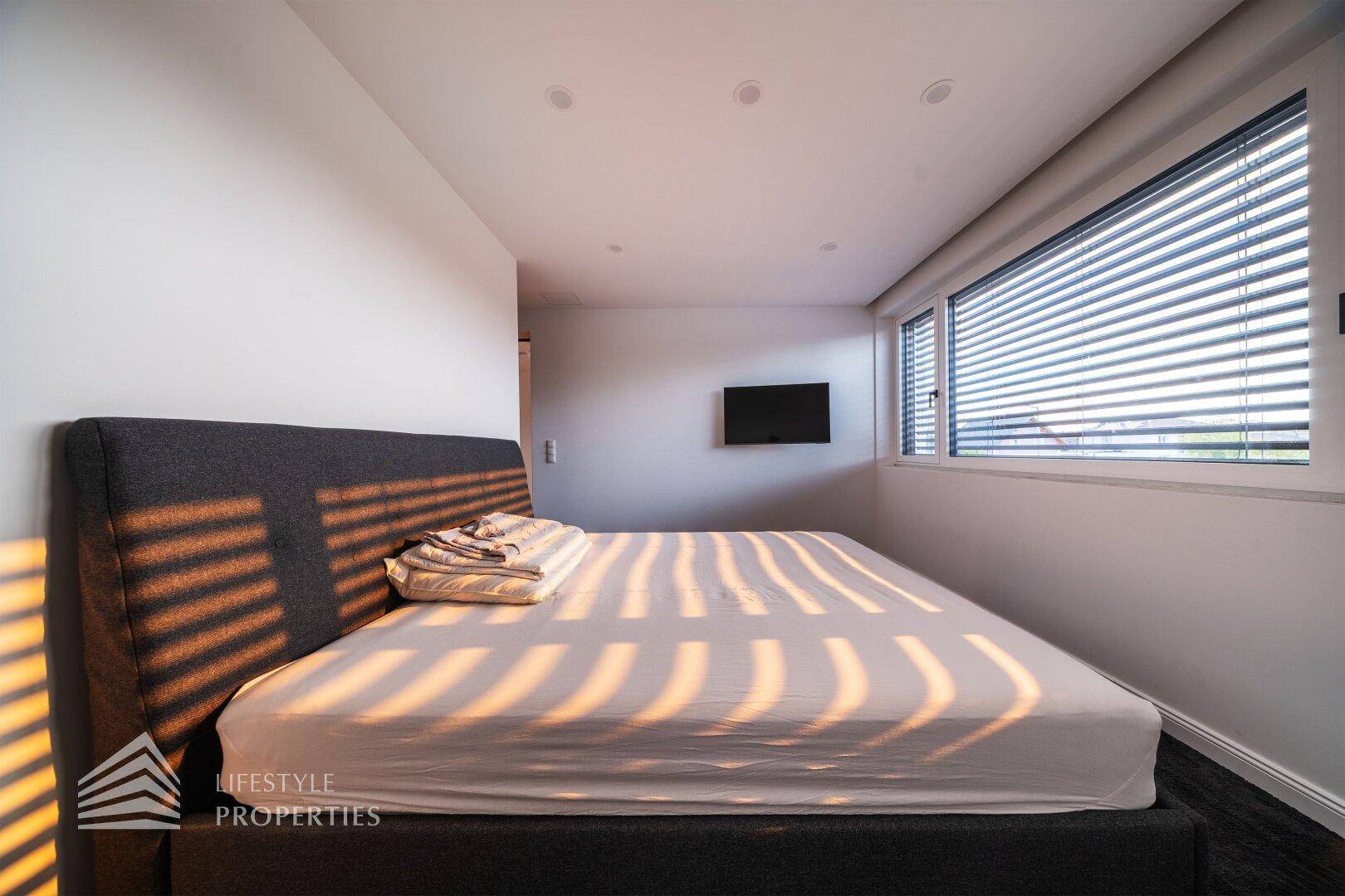 Sonnenlichtdurchflutetes Schlafzimmer mit modernem Bett und Jalousien am Fenster.