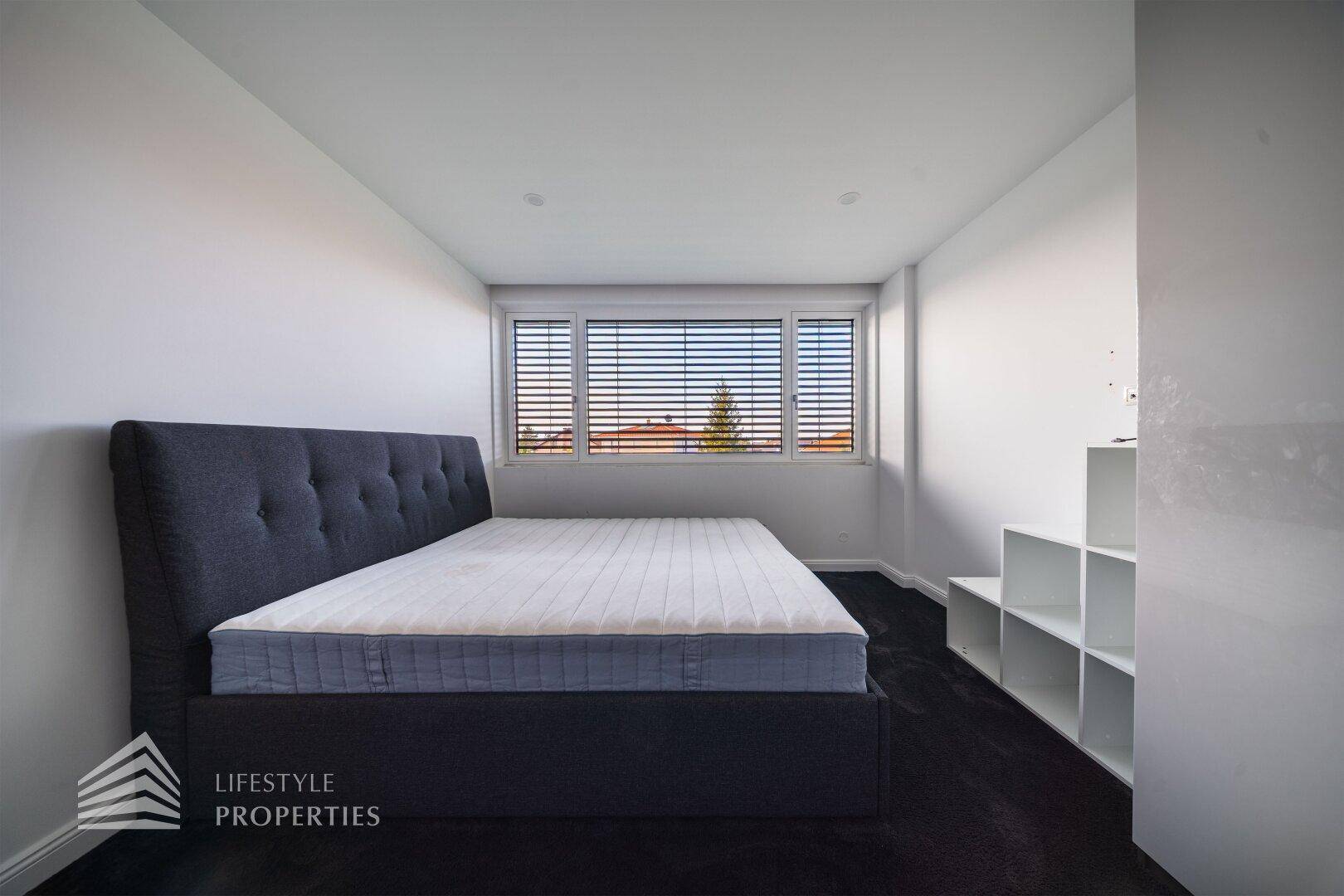 Modernes Schlafzimmer mit dunklem Teppichboden, großem Bett und Fenster mit Jalousien.