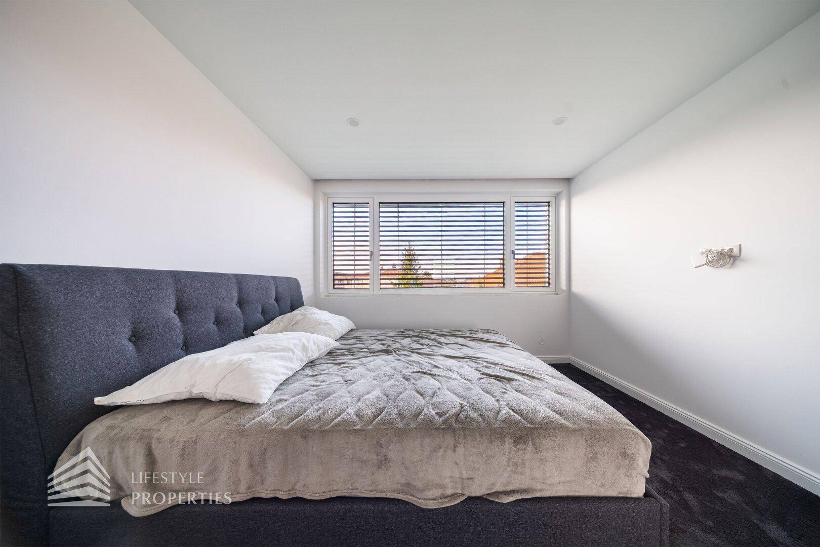 Helles Schlafzimmer mit minimalistischer Einrichtung, großem Bett und Fenster mit Jalousien.