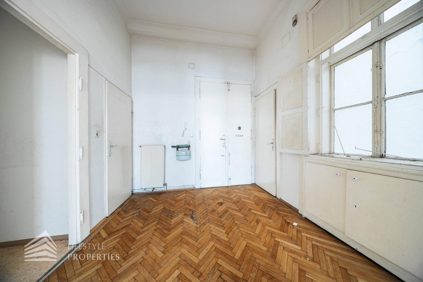 Leerer Raum mit Fischgrätparkett, Fenster und Einbauschränken, der eine umfassende Renovierung benötigt.