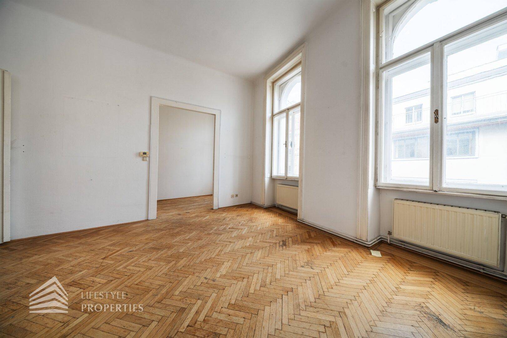 Geräumiges Zimmer mit Fischgrätparkett, hohen Fenstern und Heizkörpern, ideal für eine Neugestaltung.